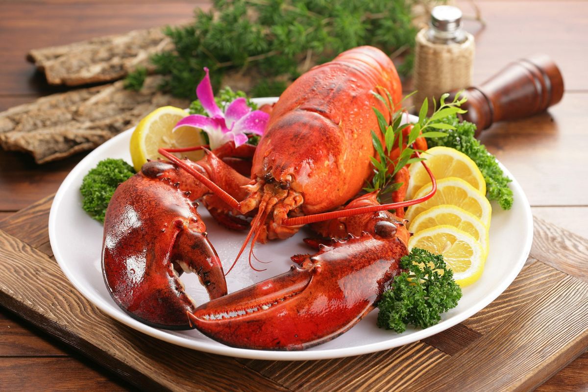 Cooked red lobster on a white plate garnished with lemon slices, parsley, and an orchid, served on a wooden table with bread in the background