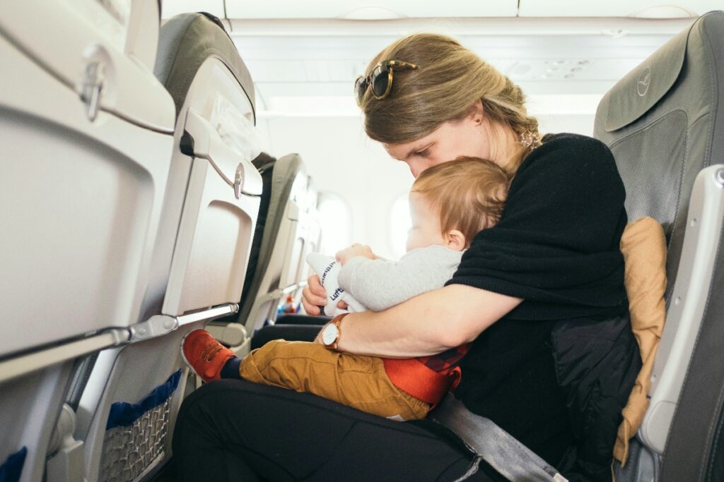 Mother in a black top sits in an airplane seat, cradling a baby while wearing a brown watch, with seats and a child’s shoe visible nearby.