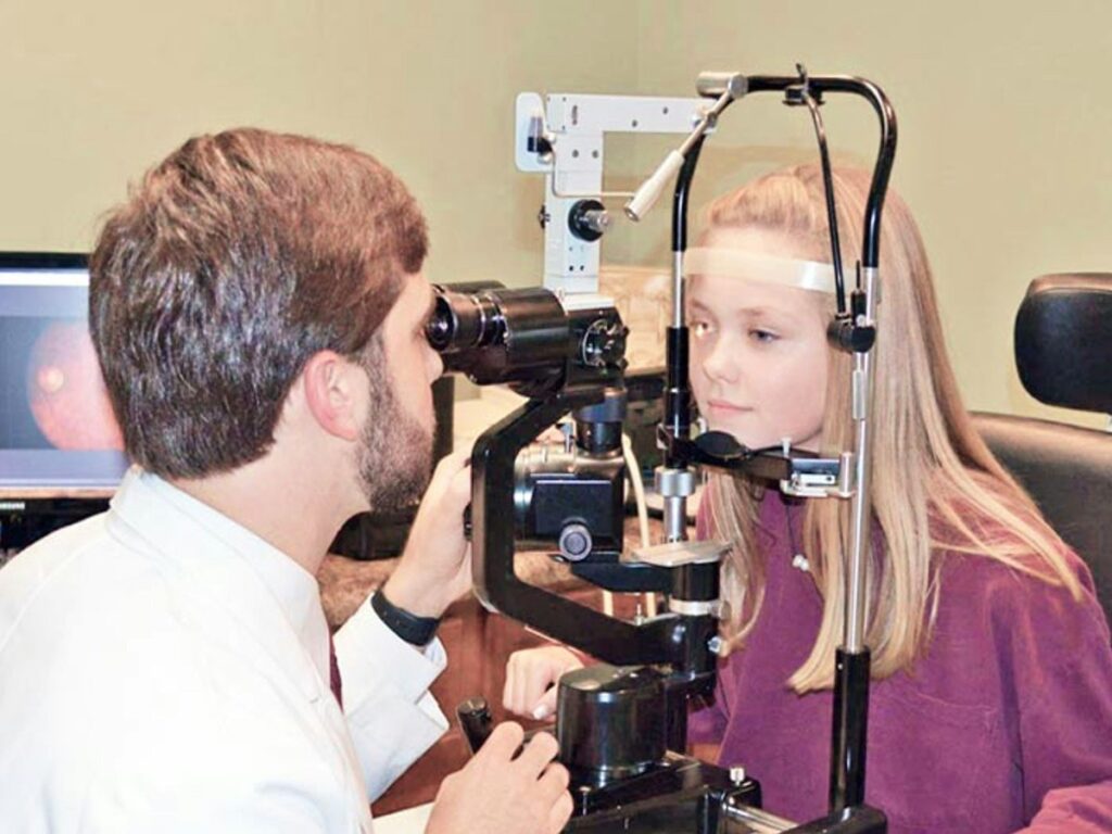 Optometrist performs eye exam on girl using slit-lamp microscope.",