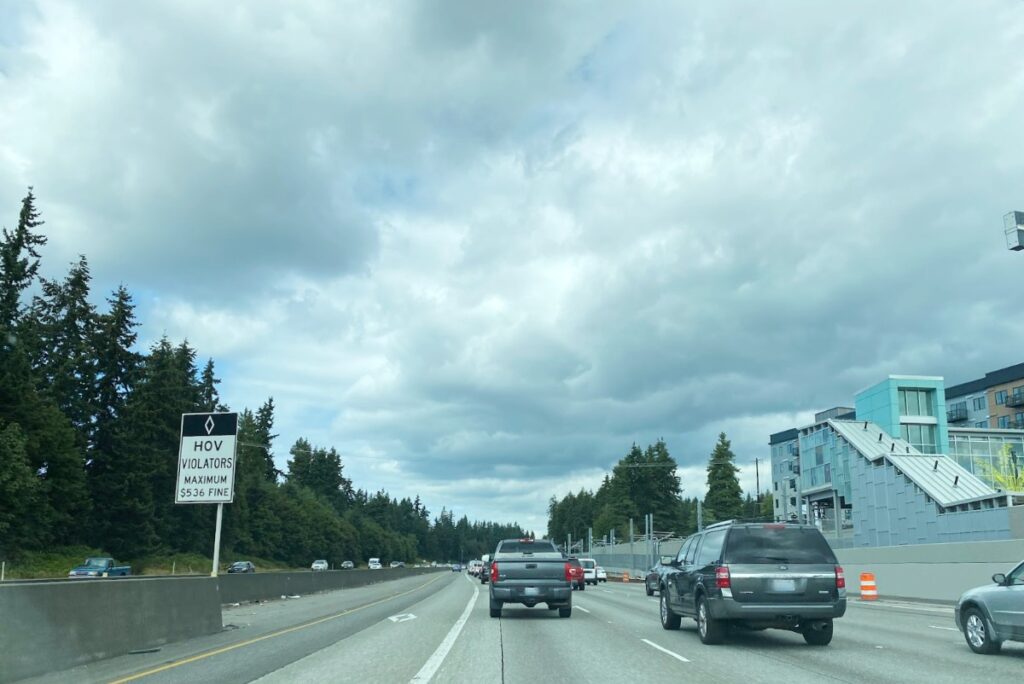 Highway scene with cars in traffic; a sign reads 'HOV Violators Maximum $536 Fine' on the left, cloudy skies above trees and a modern building on the right