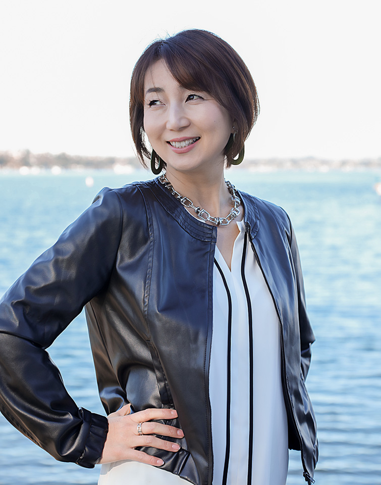 Portrait of a smiling woman by the water, wearing a navy leather jacket and silver jewelry, looking off to the side