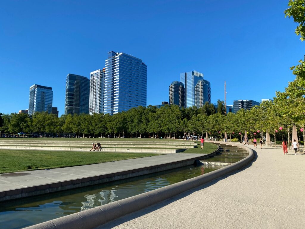 bellevue-15 - junglecity.com Curved gravel path by a reflective canal in a city park with green trees and tall glass skyscrapers in the background under a blue sky, people strolling.