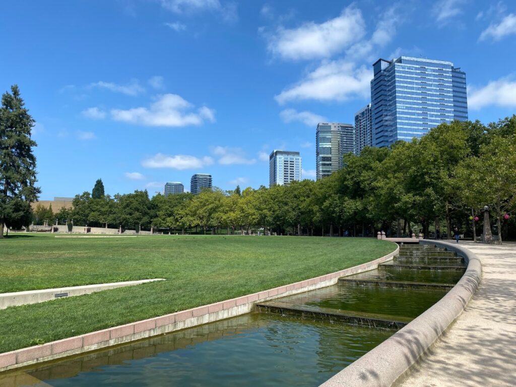bellevue-30 - junglecity.com Wide city park with a curved concrete canal and a grassy field, tall modern buildings in the background under a blue sky.