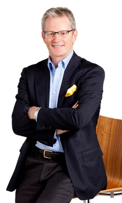 Businessman in a dark blazer and light blue shirt, wearing glasses and a yellow pocket square, standing with arms crossed beside a wooden chair.