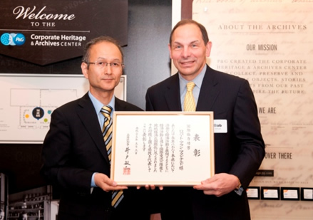 Two men in suits pose holding a framed certificate in front of archive center signage.