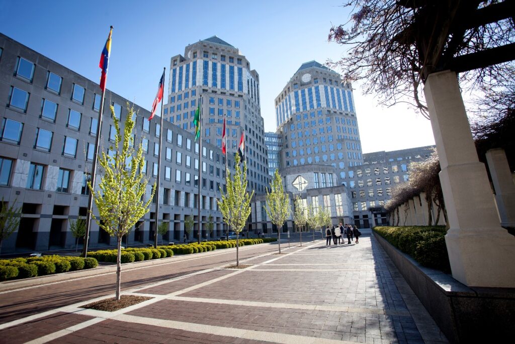 Wide view of a modern office complex with flagpoles, trees, and a group of people walking along a brick-pathed plaza on a sunny day in front of tall blue-tinted buildings.