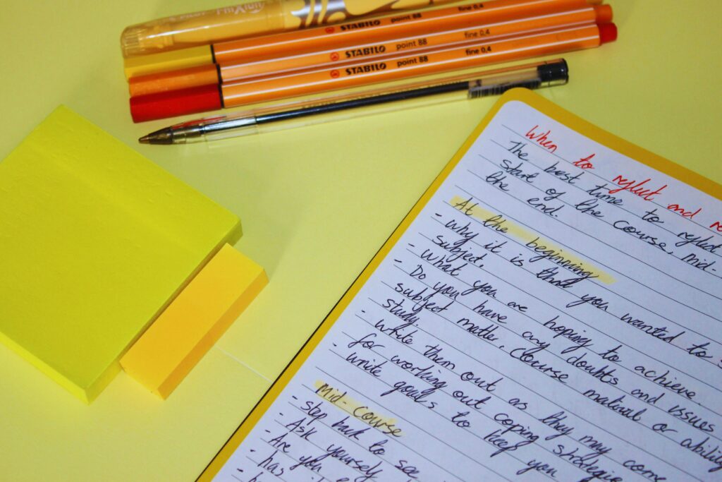 Yellow desk with stacked sticky notes, markers, and a handwritten to-do list on a yellow notebook page nearby.