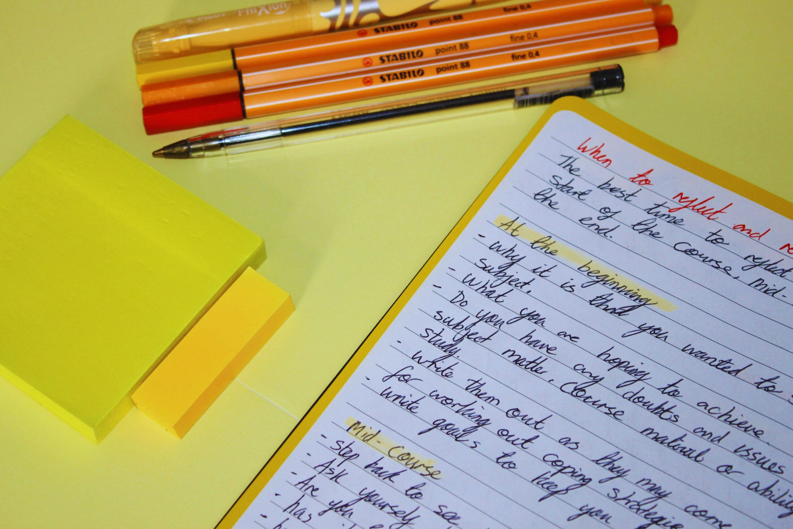 Yellow desk with stacked sticky notes, markers, and a handwritten to-do list on a yellow notebook page nearby.