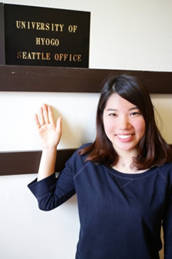 essay-hyogo-4-1 - junglecity.com Smiling woman waves beside a sign that reads 'University of Hyogo Seattle Office'.