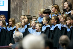essay-hyogo-4-7 - junglecity.com Children's choir wearing black robes with blue collars, singing on a stage with a few adults at the back; audience blurred in the foreground.