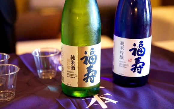 Two sake bottles (green and blue) on a purple tablecloth with small cups nearby; labels show Japanese characters in focus.