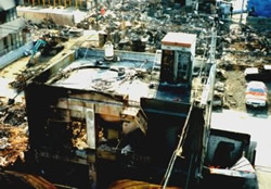 Charred, collapsed building and rubble in a scorched urban area after a fire, with damaged vehicles and extensive debris.