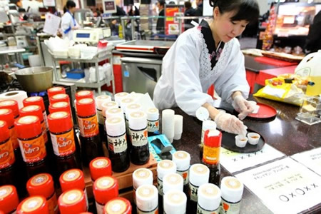 Female sushi chef rolling a sushi roll at a busy market counter with sauce bottles in the foreground