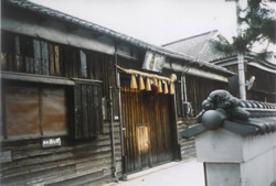 Row of dark wooden shopfronts with hanging noren curtains, beside a stone wall and decorative tiled gateway.