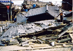 Street scene with collapsed buildings and rubble after a disaster, showing debris and damaged roofs in the foreground and background.