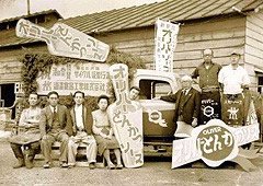 Vintage sepia photo of a group of people posing in front of a wooden building, holding large Japanese banners and signs; a truck is parked nearby.