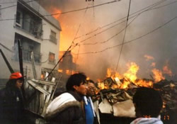 People stand in a street watching a nearby apartment building on fire with bright flames and thick smoke over the scene.