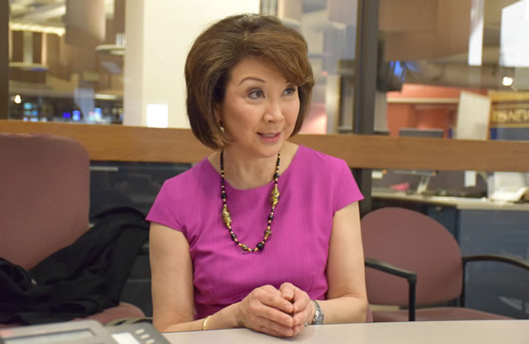 Woman in a magenta top sits at a desk in an office, hands folded, looking to her right.