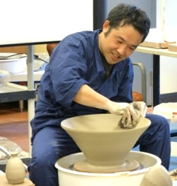 Potter shaping a clay bowl on a spinning pottery wheel, wearing a blue smock in a studio setting.