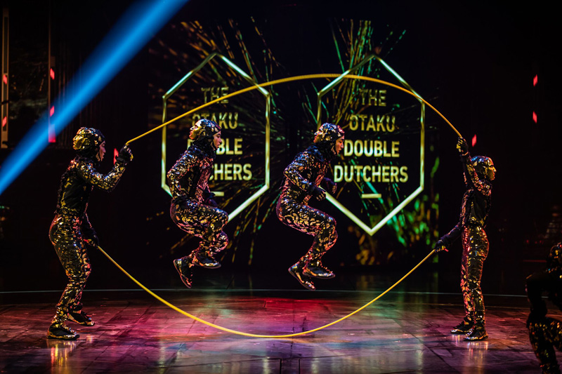 Four performers in mirrored suits jump through a glowing yellow hoop on a dark stage with neon shapes in the background.