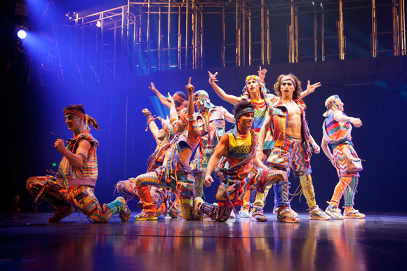 A group of dancers in bright striped outfits performing a coordinated routine on a stage under blue lighting.