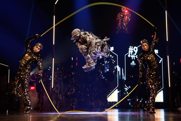 Three dancers in sequined outfits perform on a stage; a silver-clad dancer leaps midair through a large yellow hoop as two others hold the hoop.