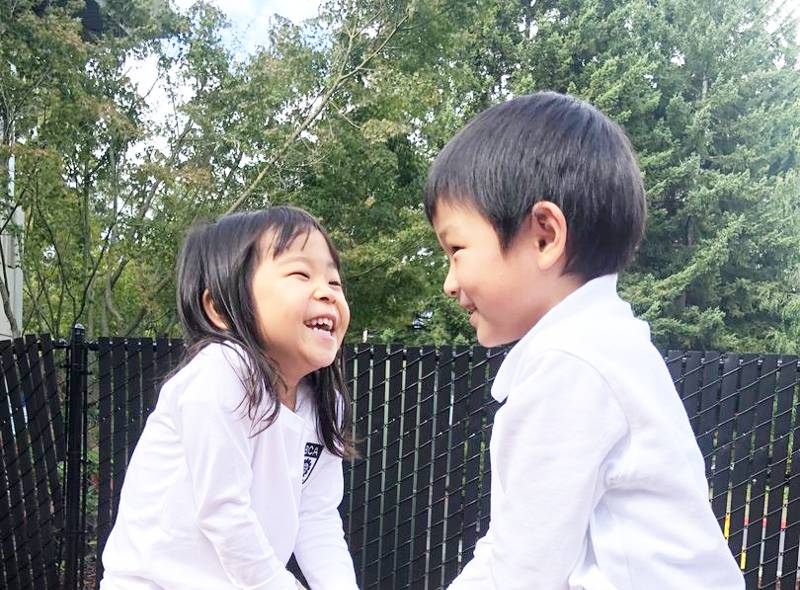 Two laughing children face each other outdoors, wearing white shirts, beside a black fence with trees in the background.
