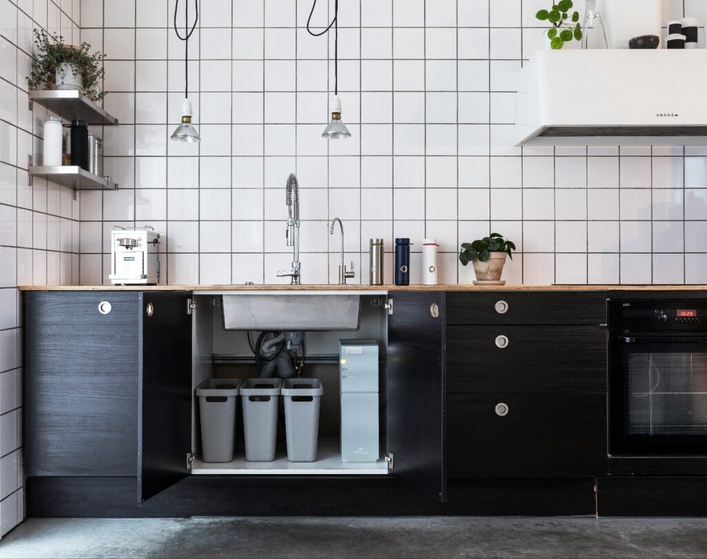 Open under-sink cabinet reveals three gray trash/recycling bins and a slim water filtration unit in a modern dark-wood kitchen cabinet.