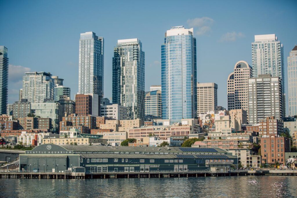 Seattle waterfront with the Seattle Aquarium in the foreground and a glass-skyscraper skyline in the background on a sunny day.