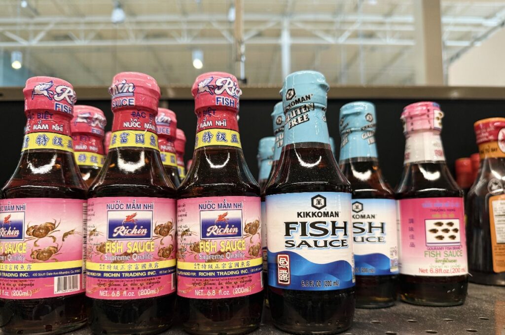 Row of fish sauce bottles on a store shelf, pink and blue labels with 'FISH SAUCE' visible on some bottles