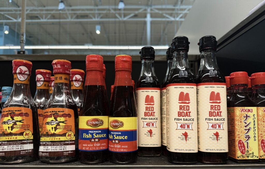 Assorted fish sauce bottles on a store shelf, including Dynasty and Red Boat brands with red caps and beige labels.