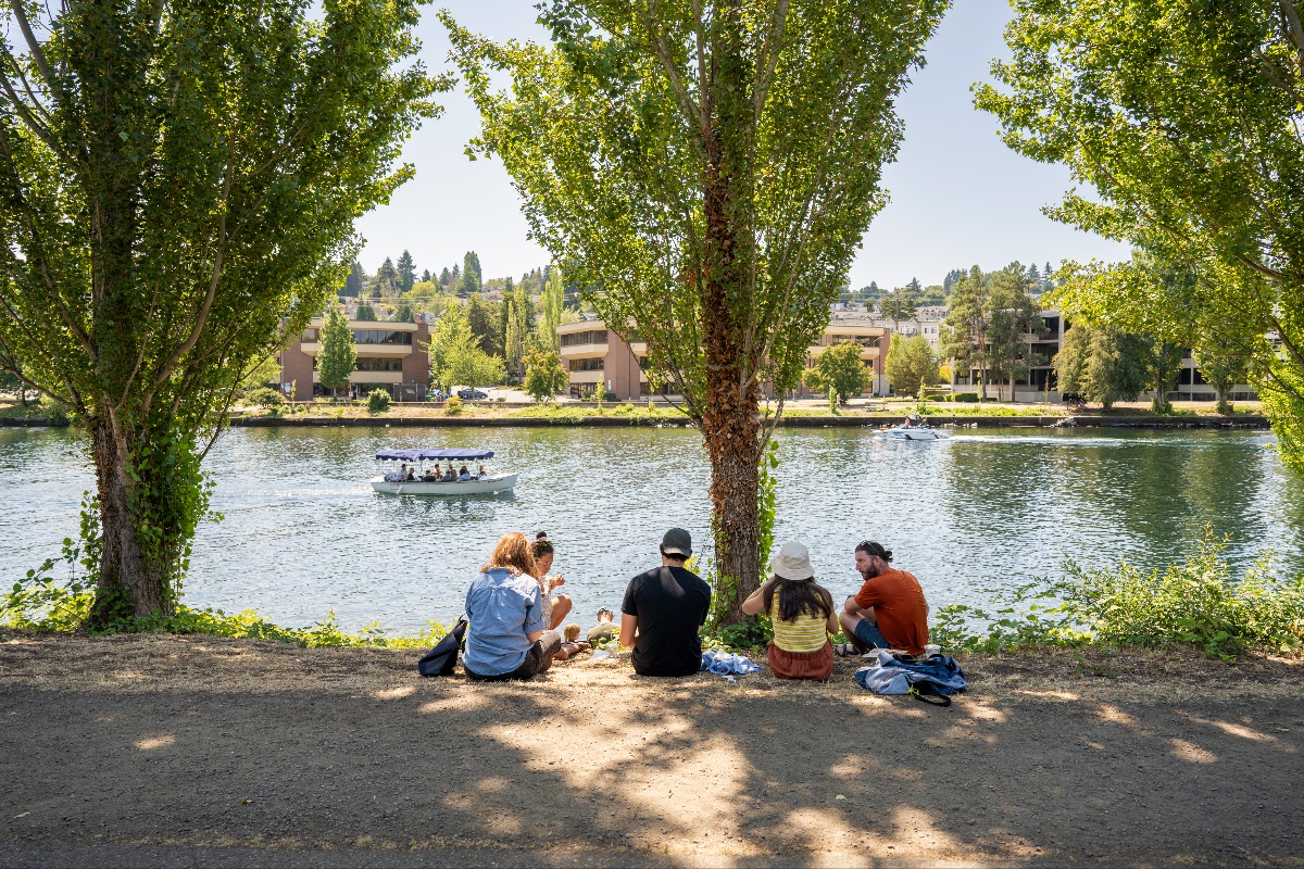 fremont-market-3 - junglecity.com Group of friends picnicking on a dirt riverside path under trees, with a boat on the water and buildings across the river.