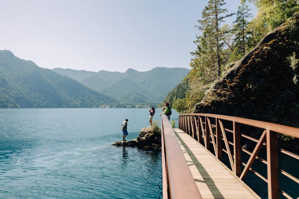 Wooden bridge over a calm blue lake with forested mountains; people on rocks taking photos nearby.