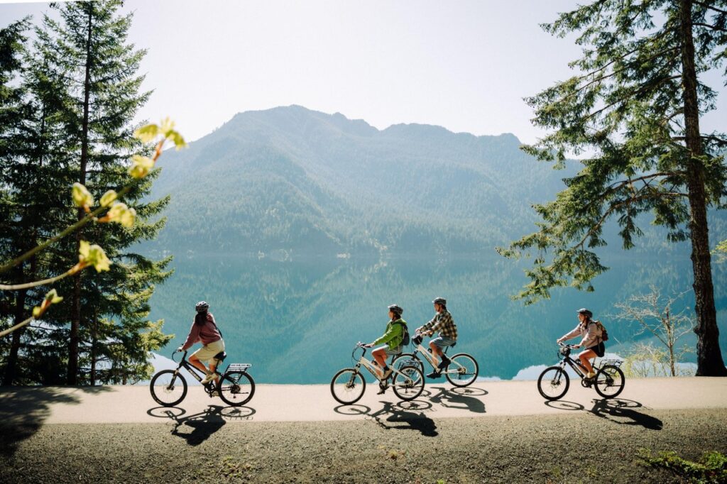 lake-crescent-6 - junglecity.com Four cyclists ride along a lakeside road with turquoise water and forested mountains in the background near tall pines.