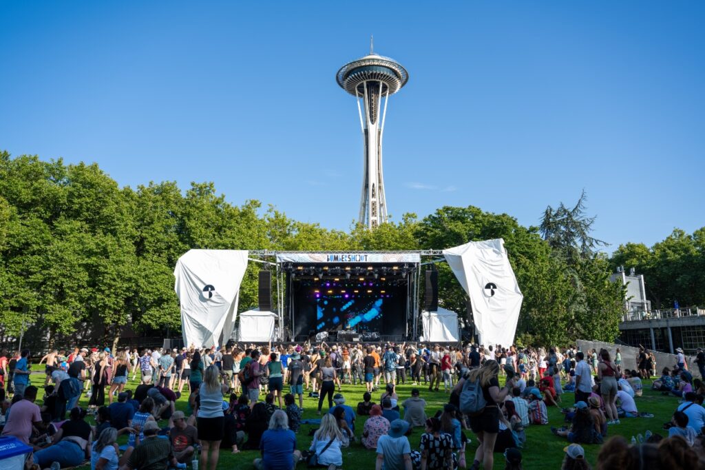 Outdoor music festival in a park with a stage, crowd, and the Space Needle in the background.