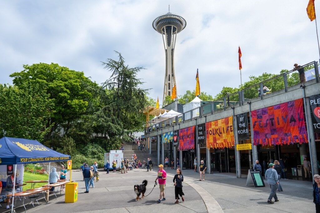 Outdoor festival at Seattle Center with colorful banners, tents, and the Space Needle in the background, sunny day.