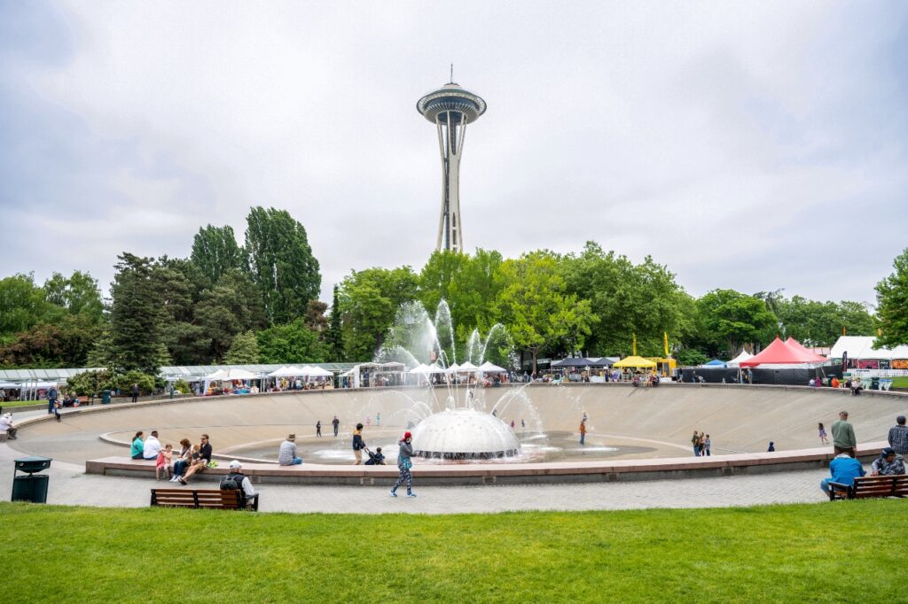 Space Needle rising behind a large circular fountain in a park, with families and vendors along the walkway around the water edge.