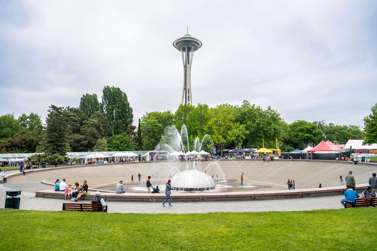 Space Needle rising behind a large circular fountain in a park, with families and vendors along the walkway around the water edge.