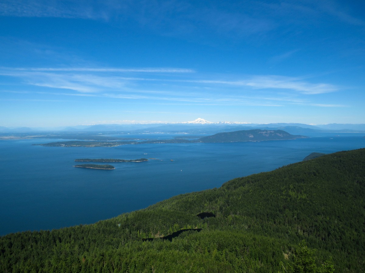 Panoramic view of a blue sea with small forested islands, a green hillside in the foreground, and snow-capped mountains on the horizon under a clear sky.