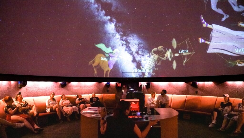 People seated around a circular sofa watching a starfield projection on a curved dome; constellations Sagittarius, Scorpio, and Libra are visible on the screen.