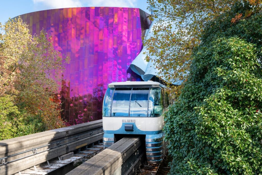 White and blue monorail train on elevated tracks beside a vibrant purple metallic wall, with trees on both sides.
