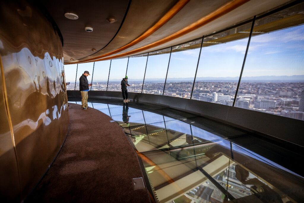 People stand at an observation deck with a glass floor, overlooking a city skyline below under a clear sky.