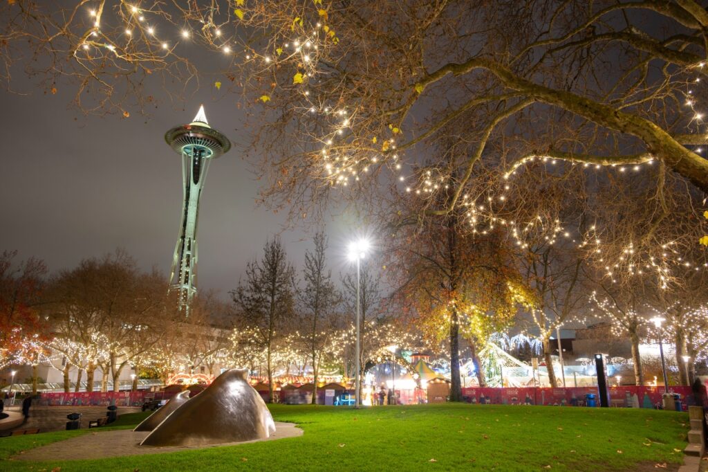 Night scene in a park with trees wrapped in string lights and the Space Needle in the distance at dusk/nightfall.