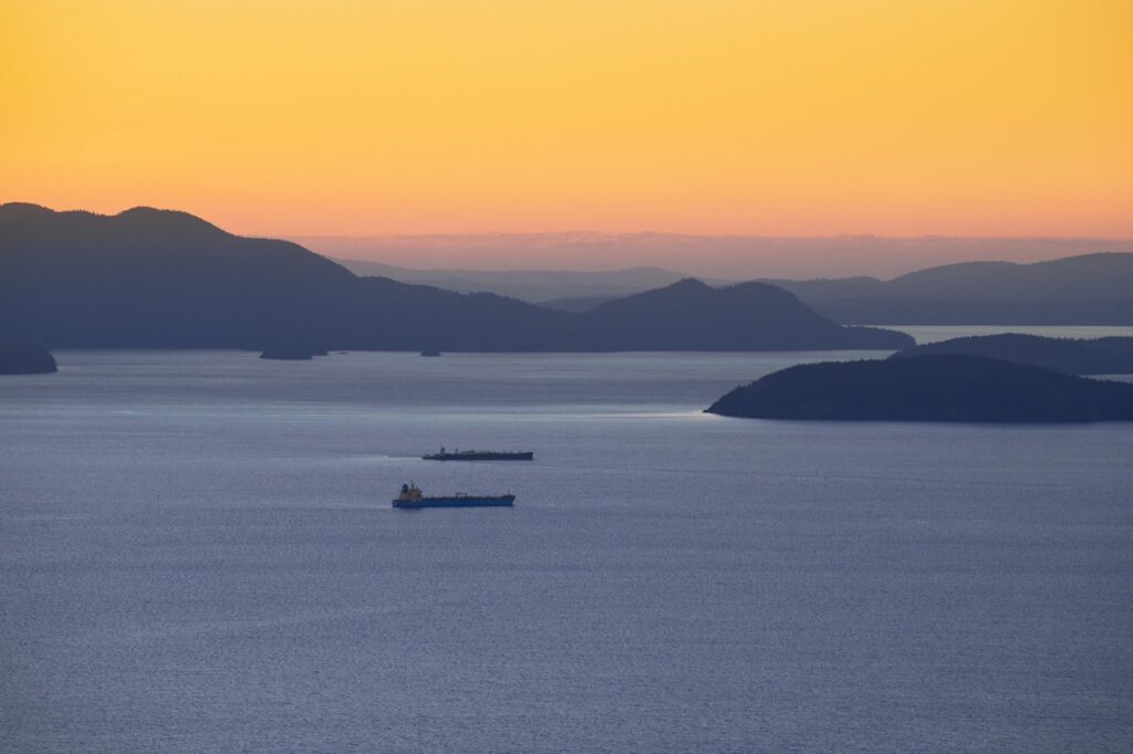 Orange sunset over a calm sea with layered silhouetted islands and two cargo ships in the foreground.