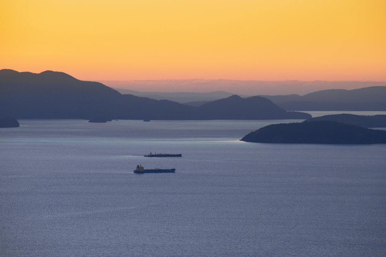 Orange sunset over a calm sea with layered silhouetted islands and two cargo ships in the foreground.