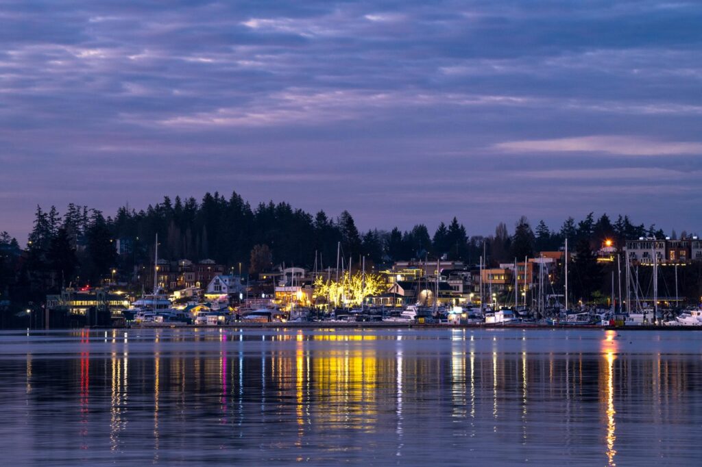 Evening harbor with boats docked along a waterfront town; warm lights reflect on calm water nearby.