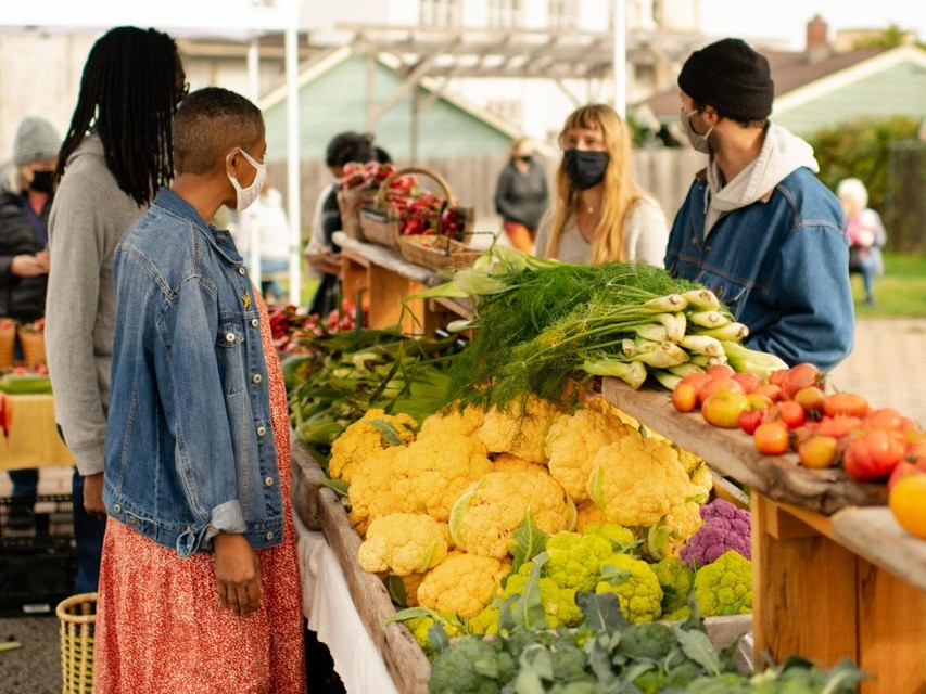 People at an outdoor farmer's market wearing masks, examining colorful fresh vegetables like cauliflower, leafy greens, and tomatoes on a stall table.