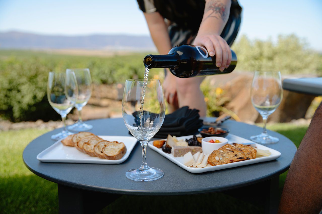 Person pours wine into a glass at an outdoor table with cheese, bread, and olives on white platters amid a vineyard setting