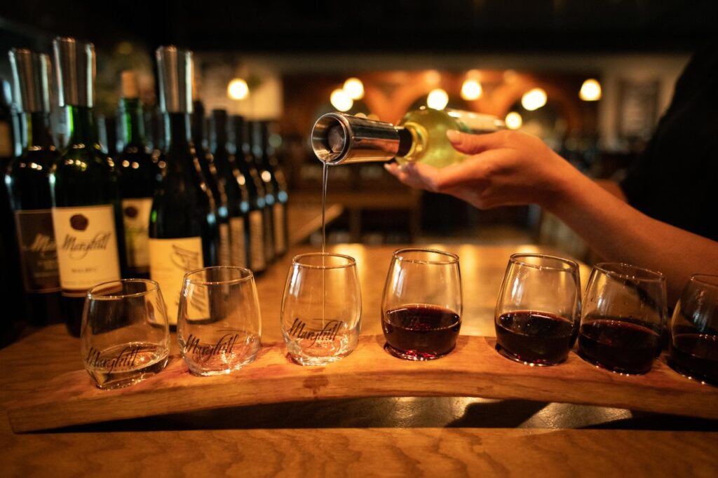 Bartender pours red wine into a row of tasting glasses on a wooden flight board at a dim bar.