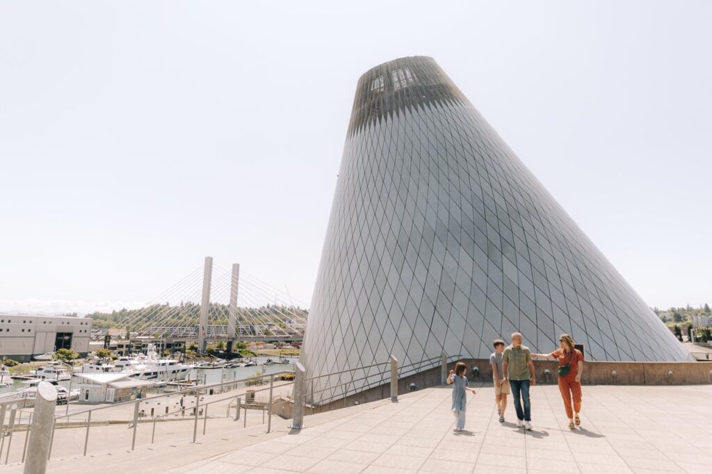 tacoma-bridge-of-glass-1 - junglecity.com Family of four walking on a sunlit terrace beside a large, cone-shaped glass building by the marina, with a bridge in the background.